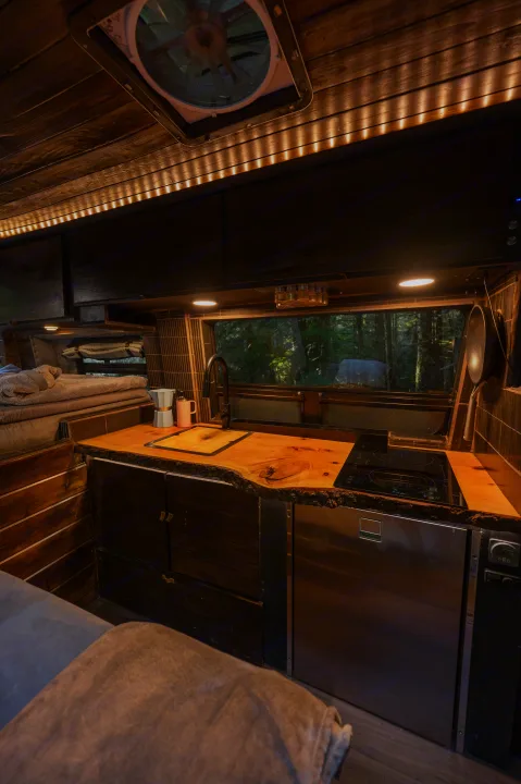 Cozy RV rental interior with a fully equipped wood kitchen, comfortable sleeping area, warm lighting, and forest views.. Mercedes-Benz Sprinter 2021