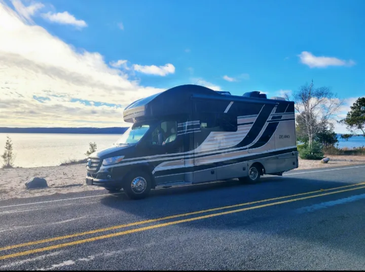 A sleek black & grey DELANO RV rental sits on a roadside, overlooking a calm lake under bright blue skies.. Thor Delano 2021