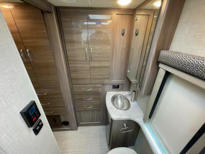 Modern RV rental bathroom with a sink, mirror, toilet (right), and extensive wood-grain cabinets and drawers (left).. Thor Delano 2021
