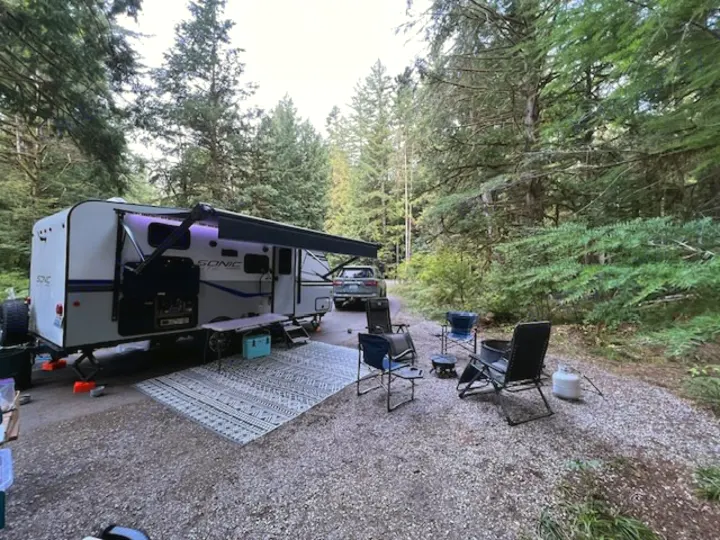 Relaxing rv rental campsite with an RV, extended awning, outdoor rug, chairs, and fire pit among tall forest trees.. Sonic Lite 2022