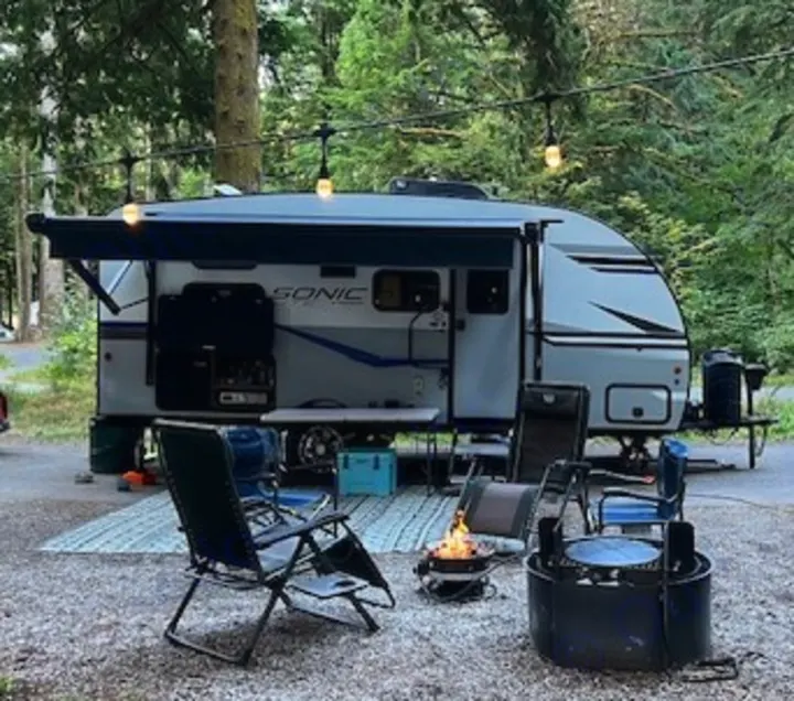 This **RV rental** makes for cozy **camping**! A light gray **travel trailer** is set up at a **campsite** with a **campfire** & chairs.. Sonic Lite 2022