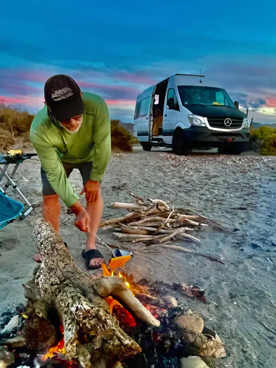 Man starting campfire by his RV rental camper van on a sandy beach at sunset, enjoying van life.. Mercedes Sprinter 2017