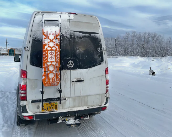 Alaska RV rental van in winter, covered in snow with traction boards, parked in a snowy landscape with a dog nearby.. Mercedes Sprinter 2017