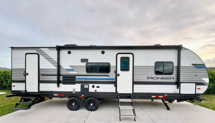 A modern gray and white travel trailer with blue accents, ideal for an RV rental adventure. Steps extended, ready for camping.. Heartland RVs Pioneer 2021
