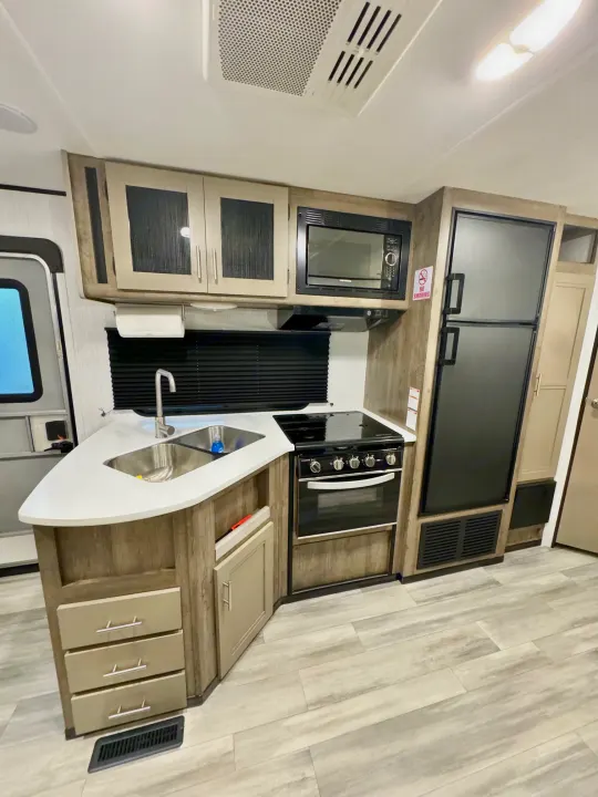 Modern RV rental kitchen featuring a double sink, black stove, microwave, refrigerator, light wood cabinets, and light flooring.. Heartland RVs Pioneer 2021