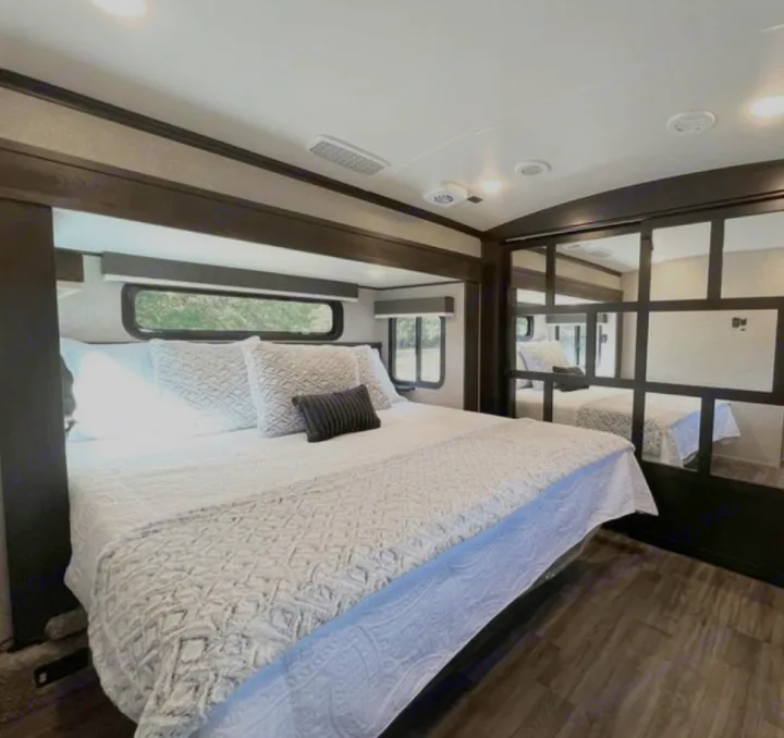 Relax in this spacious RV rental bedroom featuring a large bed with white bedding, windows, & mirrored closet. Ideal for travel.. Jayco North Point 2023