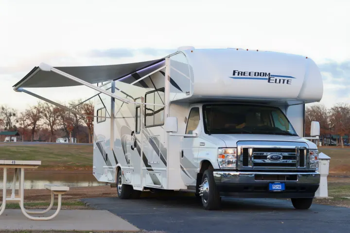White Freedom Elite `rv rental` motorhome with extended awning, parked at a lakeside campsite, perfect for camping.. Thor Freedom Elite 2023