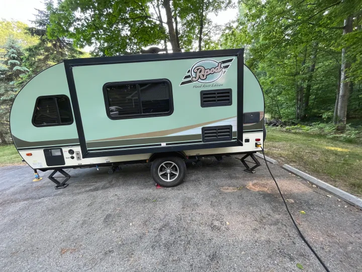A green R-Pod travel trailer is set up with stabilizer jacks in a tree-lined campground, ready for an RV rental adventure.. R-Pod Hood River Edition 2016