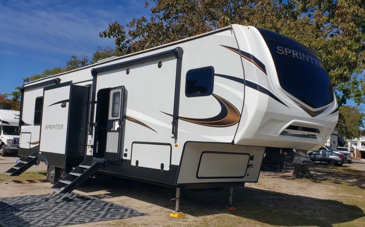 A modern white Sprinter fifth-wheel RV rental with multiple slide-outs deployed, set up in a bright, tree-lined park.. Keystone Sprinter 2022