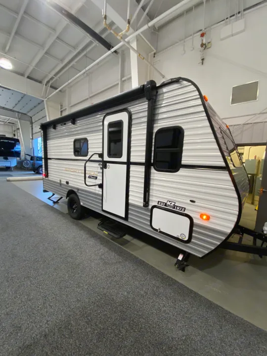 Side view of a white & grey KZ travel trailer for rv rental, with a door, windows, and rolled-up awning, parked indoors on carpet.. KZ Sportsmen Classic 180BH 2024