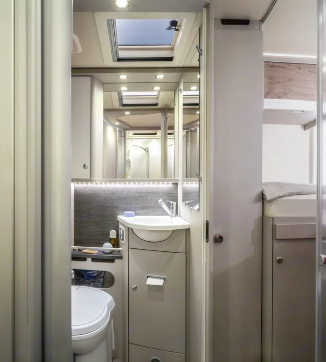 Inside a modern RV rental: a compact bathroom with toilet, sink, and mirrored vanity. A comfortable bed area is also visible.. Hymer Exis I 678 2024