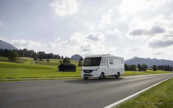 A white RV rental drives through a picturesque landscape of rolling green hills, forests, and mountains under a cloudy sky.. Hymer Exis I 678 2024
