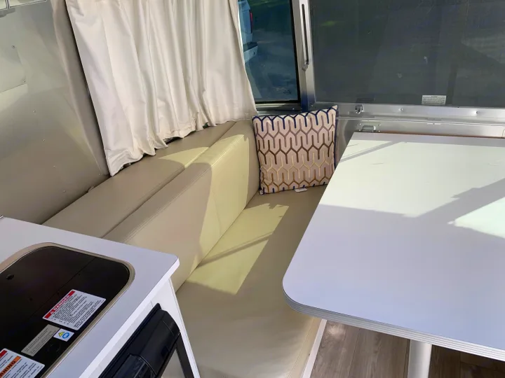 RV rental interior shows a bright dinette with cream seating, white table, and compact kitchen counter with safety warnings.. Airstream Sport 2019