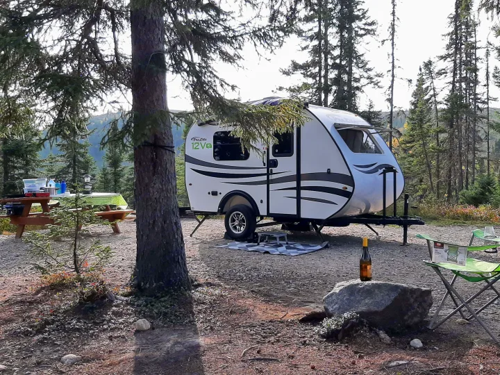 Peaceful nature escape! A compact white **rv rental** travel trailer is parked at a wooded campsite with a picnic table and green chairs.. Prolite 12 V 2019