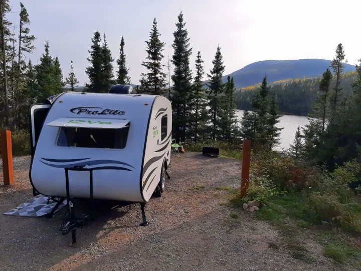 A ProLite RV rental is parked at a picturesque lakeside campsite, featuring a travel trailer amidst evergreen trees and a scenic mountain backdrop.. Prolite 12 V 2019