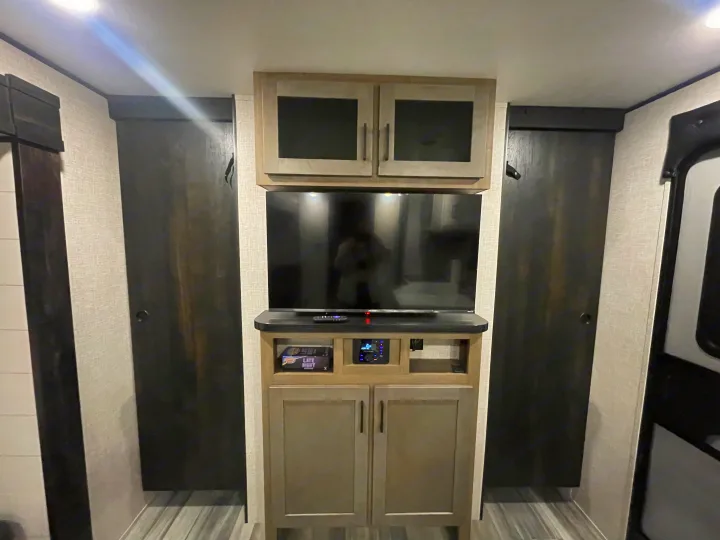 RV rental interior: A central entertainment setup with TV, media system, and various cabinets. Flanked by dark wardrobe doors.. Jayco Jay Flight 261BHS 2024