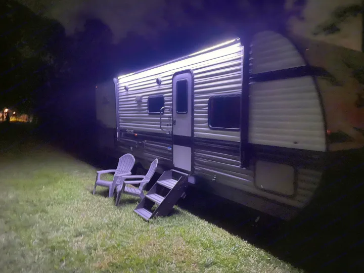 An illuminated RV rental at night, parked on green grass. Two white Adirondack chairs and steps offer a welcoming outdoor leisure space.. Jayco Jay Flight 261BHS 2024