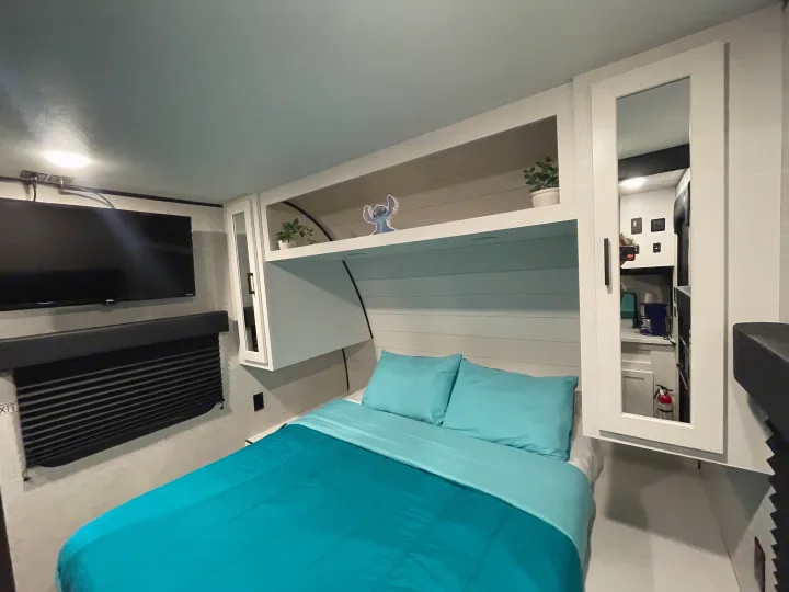 Step into this modern RV rental bedroom, featuring a comfy teal bed, TV, clever overhead storage, and a reflected kitchen view.. Jayco Jay Flight 261BHS 2024