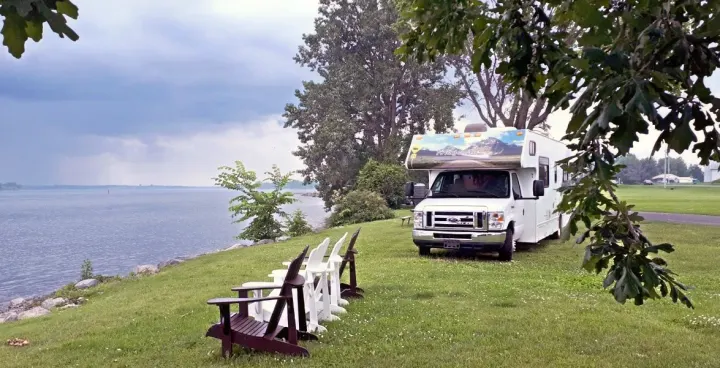 An RV rental is parked by a tranquil lake, with Adirondack chairs facing the water, offering a peaceful lakeside retreat.. Thor Majestic 2018