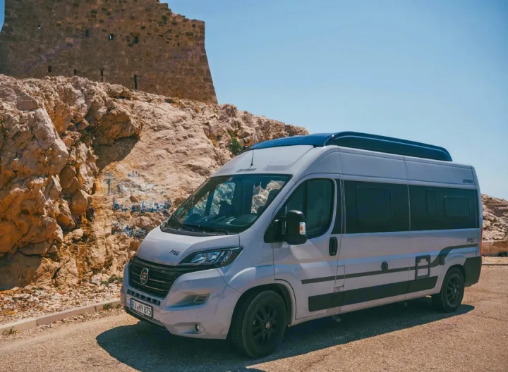 RV rental campervan near a historic stone fortress. Light gray van parked on a rocky path under a clear blue sky.. Hymer FREE 600 CAMPUS 2024