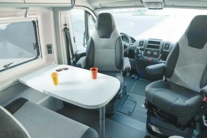 Inside an RV rental: a bright interior with a white dining table holding two cups and sunglasses, overlooking the vehicle's front seats.. Hymer FREE 600 CAMPUS 2024