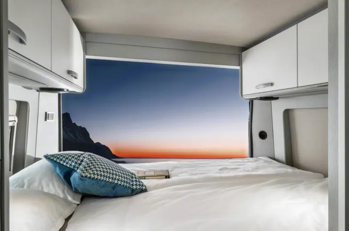 Inside a comfortable RV rental bedroom, a pristine bed faces a breathtaking mountain and sunset vista.. Hymer FREE 600 CAMPUS 2024