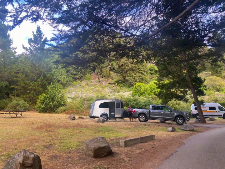RV rental campsite scene: A man with his truck and a silver teardrop camper, plus another van, are parked in a wooded area.. Airstream Basecamp 2017