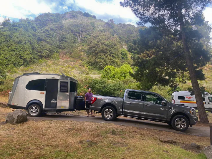 A man stands with his truck and a unique RV rental travel trailer at a beautiful forested campsite, near another camper van.. Airstream Basecamp 2017