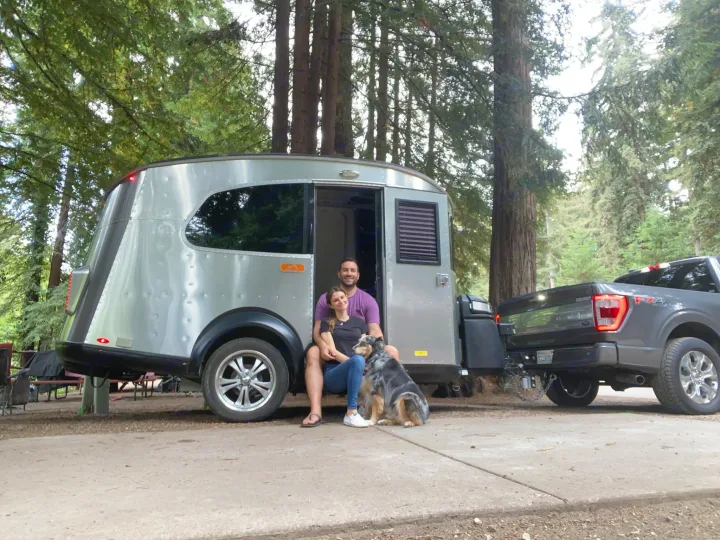 A happy couple and their dog pose by their modern silver rv rental, hitched to a truck in a scenic wooded campsite.. Airstream Basecamp 2017