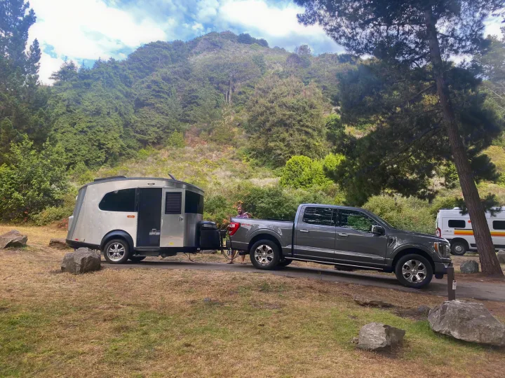 RV rental adventure! A grey pickup truck tows a silver travel trailer at a lush, forested campsite, with another camper van nearby.. Airstream Basecamp 2017