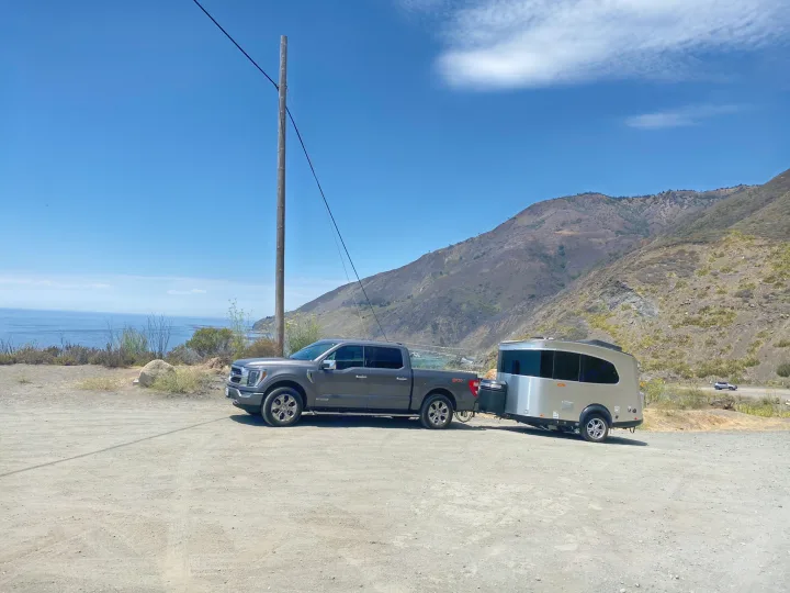 Scenic coast view with an RV rental: A pickup truck towing a sleek travel trailer on a dirt lot, mountains & ocean in the background.. Airstream Basecamp 2017