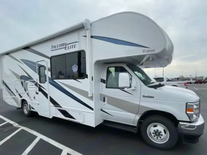 This white Class C motorhome with blue/gray graphics, branded "Freedom Elite," is parked on asphalt. Ideal for an RV rental adventure.. Thor Freedom Elite 2023