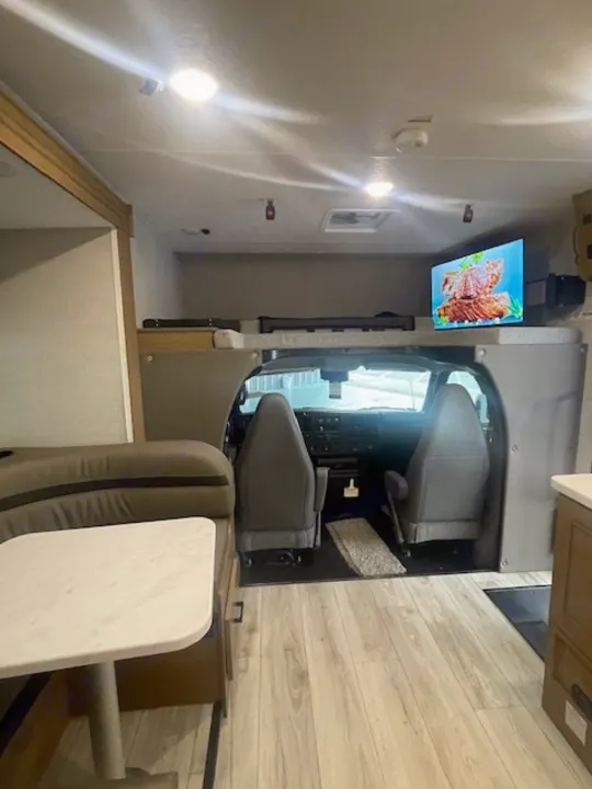 Bright RV rental interior features a dinette, overhead bunk, and cockpit with TV. Ready for comfortable adventures.. Coachmen LEPRECHAUN 230FS 2026