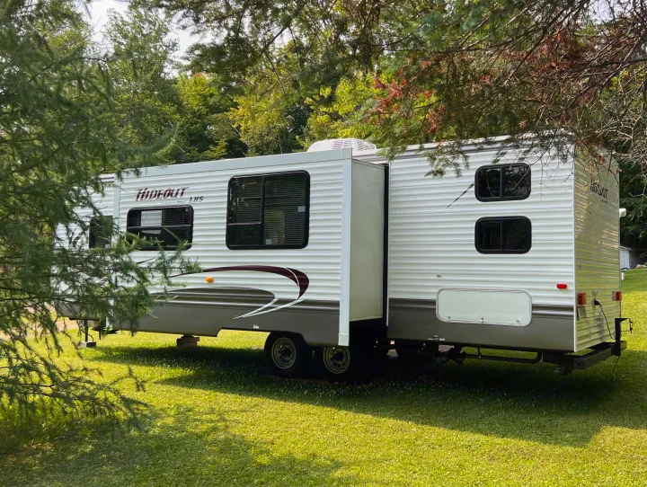 A white Hideout travel trailer, an RV rental, is parked on a sunny grassy lot surrounded by trees, with its slide-out extended.. Keystone Hideout LHS 2014