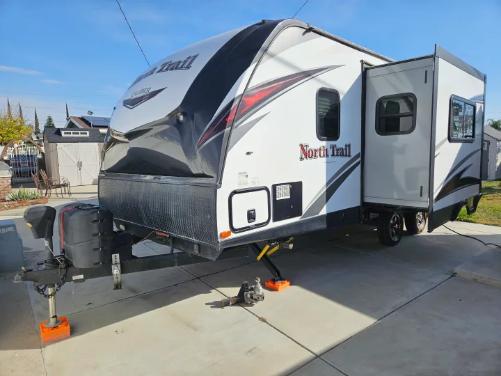A white North Trail Caliber travel trailer with an extended slide-out, parked on a concrete driveway, ready for an RV rental trip on a sunny day.. Heartland RVs North Trail 2019