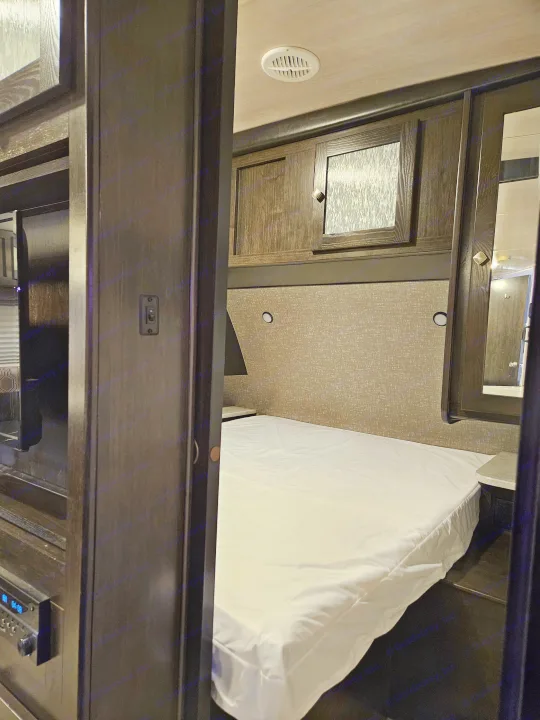 Compact RV rental bedroom with a white bed, dark wood overhead cabinets, a nightstand, and an entertainment system.. Heartland RVs North Trail 2019