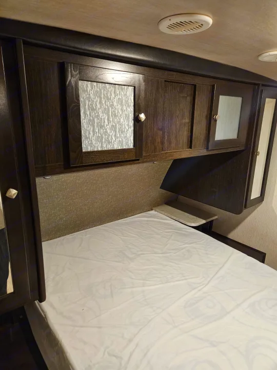 An RV rental bedroom with dark wood cabinets, a bed with white sheets, and ceiling vents, showing compact, functional design.. Heartland RVs North Trail 2019