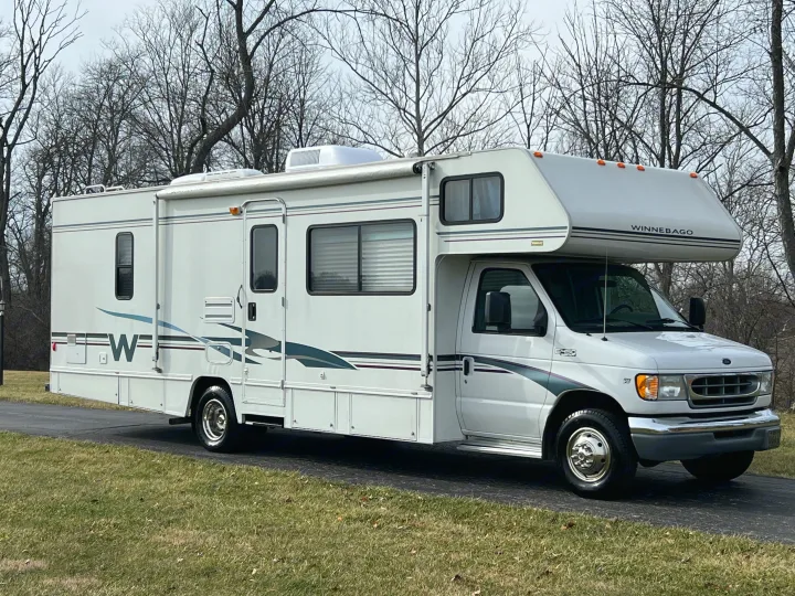 A clean white Winnebago Class C motorhome, perfect for an RV rental adventure, parked on a paved driveway with bare trees behind.. Winnebago Minnie 2002