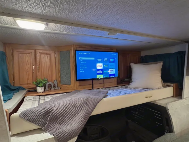 Cozy RV rental interior: a bunk bed with a gray blanket, Roku TV with streaming apps, and wood cabinets. Perfect for trips!. Winnebago Minnie 2002