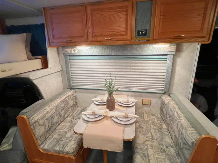 This RV rental interior showcases a tidy dining area with a set table, flanked by upholstered booth seating and wooden cabinets.. Winnebago Minnie 2002