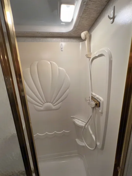 Compact RV rental shower with a seashell-themed back wall, handheld showerhead, and small shelving, perfect for travel.. Winnebago Minnie 2002
