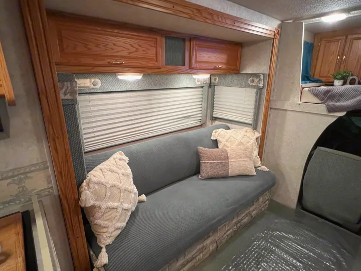 Cozy RV rental interior featuring a long upholstered bench with decorative pillows, wooden cabinets, and window blinds.. Winnebago Minnie 2002