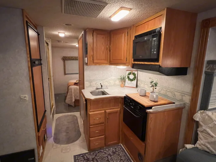 Cozy RV rental kitchen & living area with wood cabinets, sink, microwave, stove, and a peek into the bedroom.. Winnebago Minnie 2002