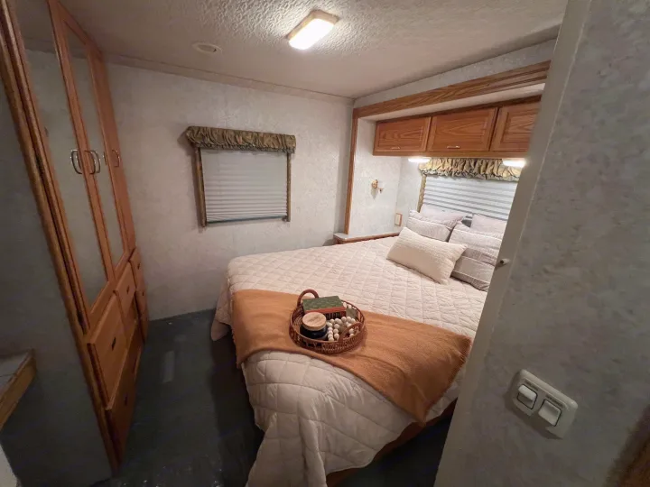Cozy RV rental bedroom with a quilted bed, pillows, window, and wooden cabinets, perfect for travel accommodations.. Winnebago Minnie 2002