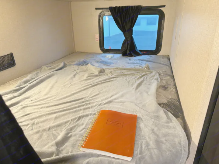 A soft grey blanket covers a cozy bunk bed inside an RV rental, with a brown notebook and an "EXIT" sign visible.. Starcraft 26BHS 2024