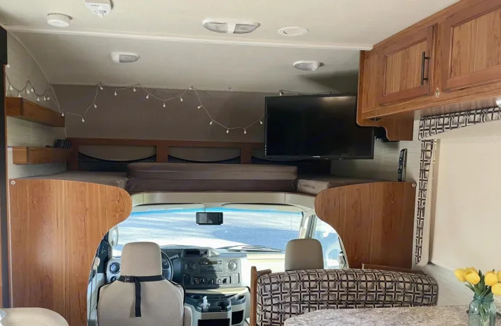 Interior of an RV rental showcasing the front cockpit, cozy over-cab bunk with string lights, and mounted TV.. Jayco Redhawk 2017