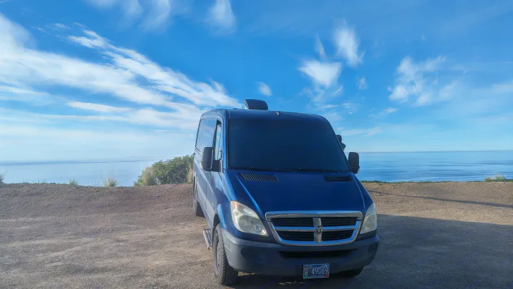 Blue RV rental van parked on a dirt cliff edge, offering stunning ocean views under a blue sky with wispy clouds.. Dodge Sprinter 2007