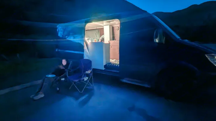 Cozy night camping in an RV rental. A person relaxes outside their illuminated camper van, set against dark mountains.. Dodge Sprinter 2007