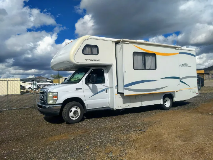 White Fleetwood Class C RV rental with an extended slide-out, parked on a gravel lot under a dramatic cloudy sky.. Fleetwood Jamboree 2009