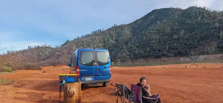 Serene lake camping with an RV rental. Blue camper van parked on red earth; person relaxing, surrounded by wooded hills.. Dodge Sprinter 2007